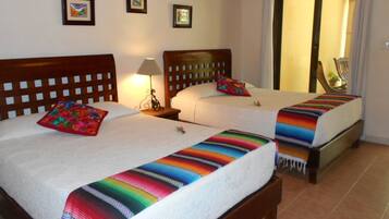 Habitación de hotel con dos camas, cada una con una manta colorida y un almohadón. Hay una mesita de noche con una lámpara entre las camas. La habitación tiene un cabecero de madera y una silla cerca de la ventana.