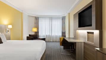 Habitación de hotel con una cama grande, un escritorio, una silla, un televisor montado en la pared y una ventana con cortinas.