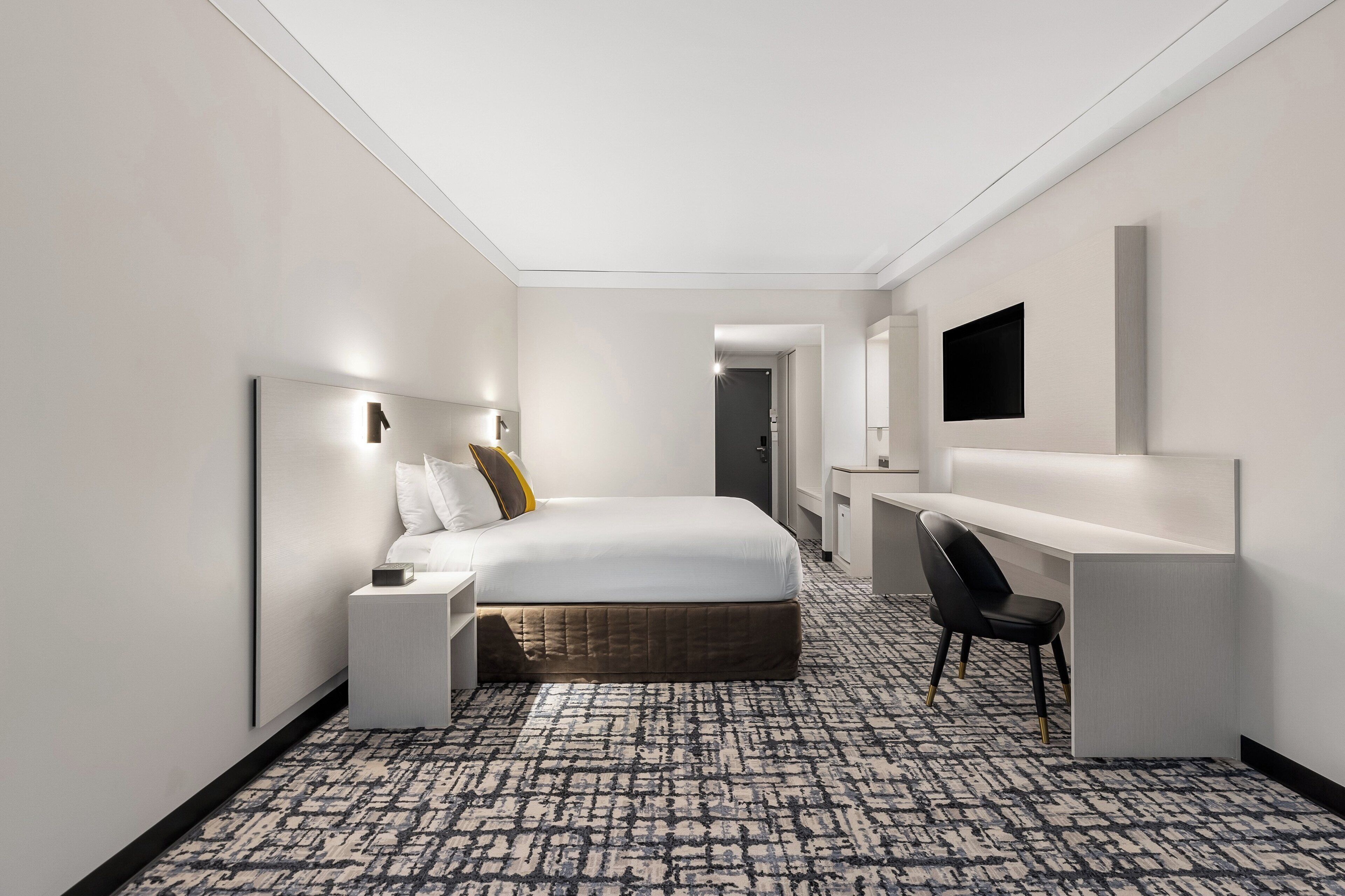A hotel room with a bed, bedside table, desk, chair, and a TV mounted on the wall.