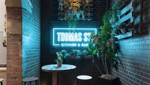 Un rincón acogedor con un letrero de neón que dice 'Thomas St. Kitchen & Bar', rodeado de paredes de ladrillo y plantas en macetas.