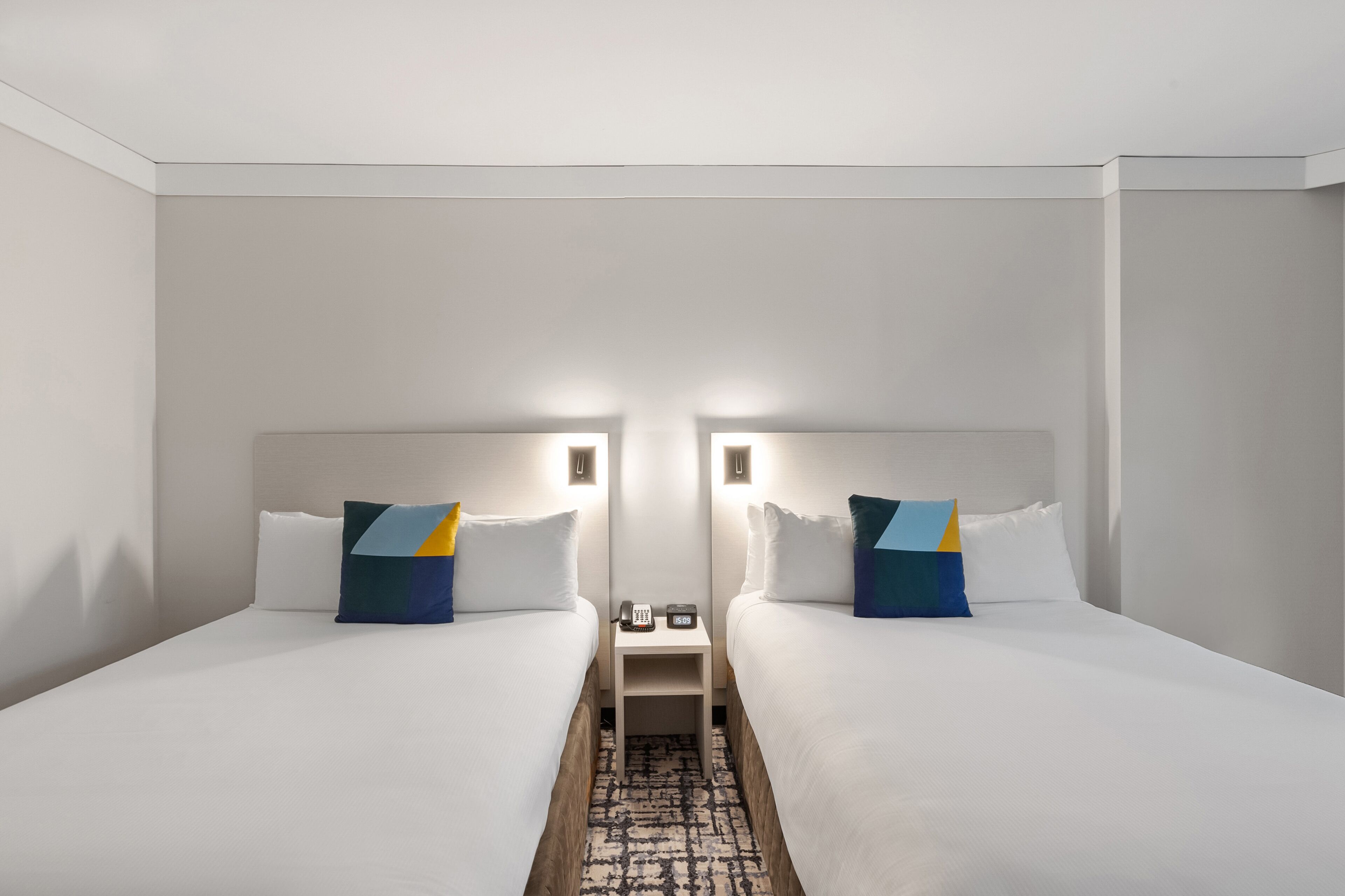 Quarto de hotel com duas camas, cada uma com cabeceira, mesa de cabeceira com telefone e abajur, e tapete estampado no chão.