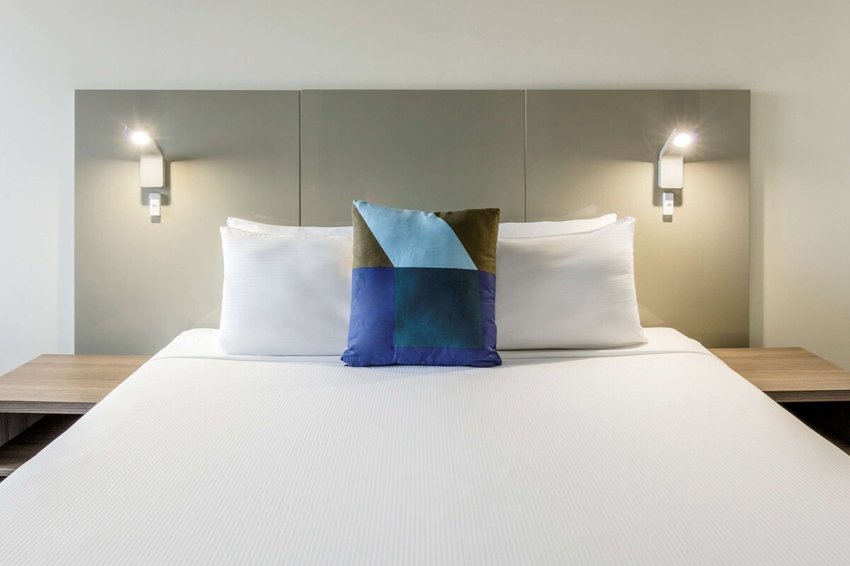A neatly made bed with two pillows, one plain and one with a geometric design, in a room with a simple headboard and wall-mounted lighting.
