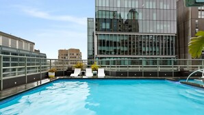 Una piscina en la azotea con barandilla de cristal, sillas de descanso blancas y un edificio moderno al fondo.