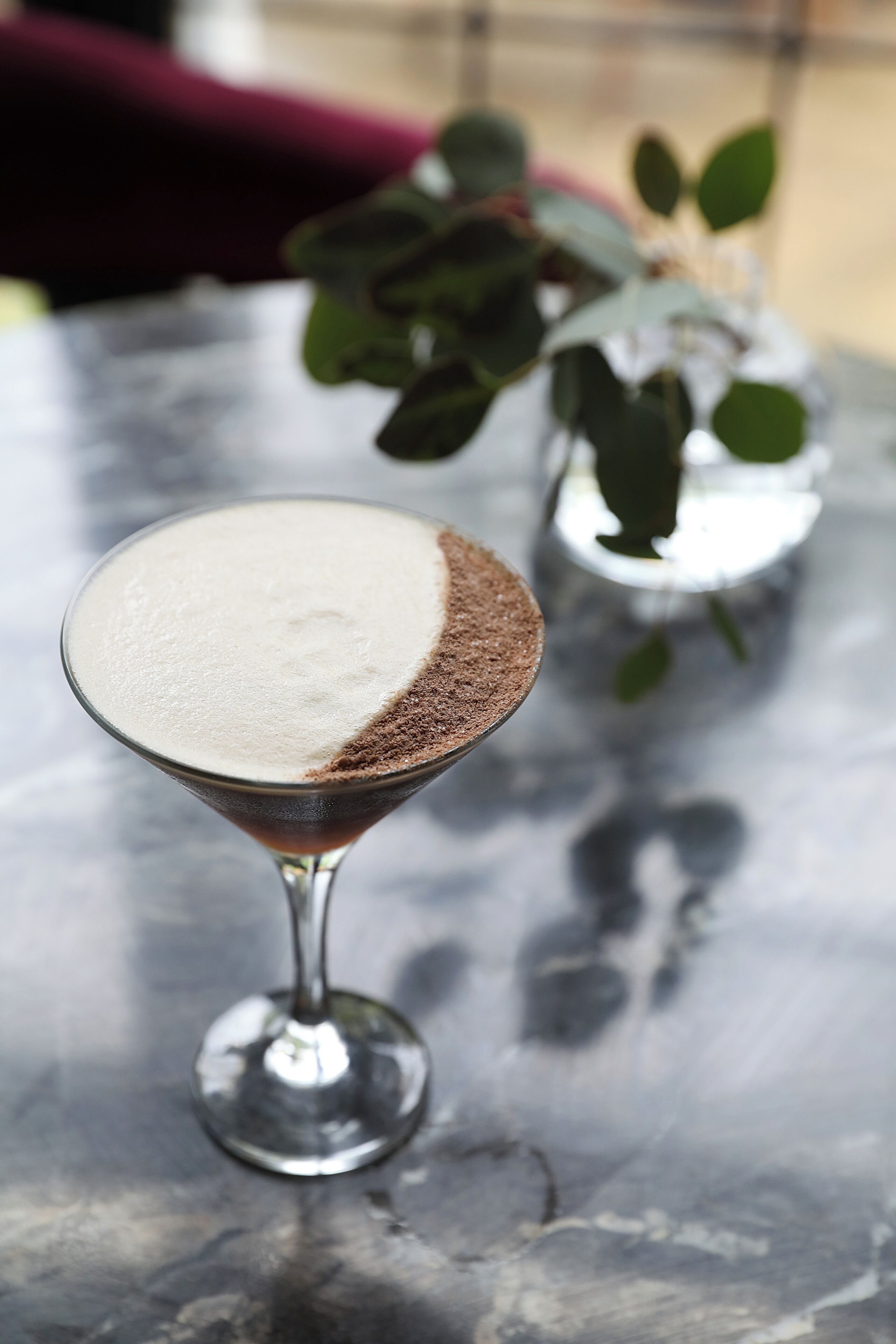 A martini glass with a frothy top and a chocolate shavings garnish on a reflective surface.