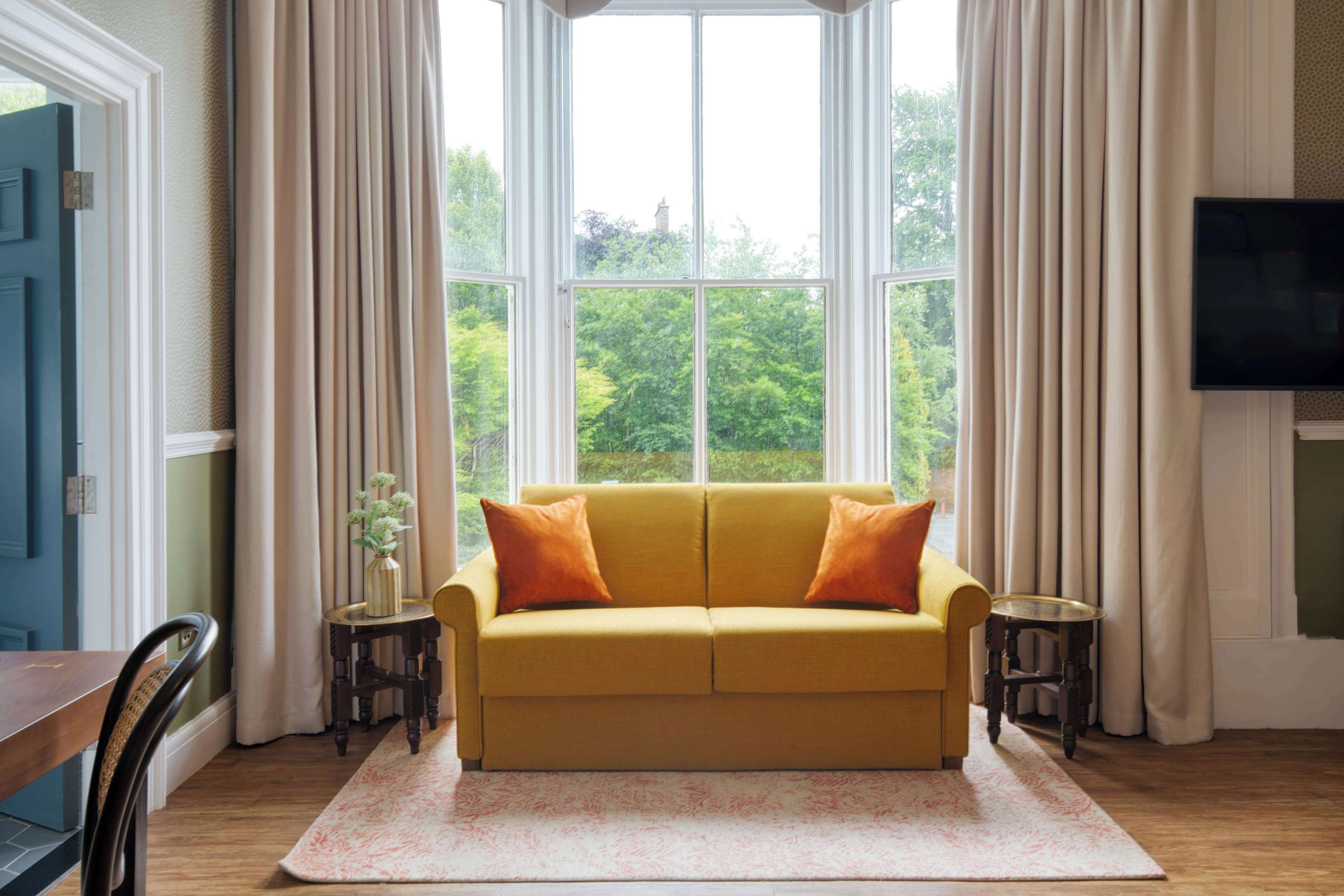 Ein Zimmer mit einem gelben Sofa, zwei Beistelltischen, einem Flachbildfernseher und einem großen Fenster mit Vorhängen.