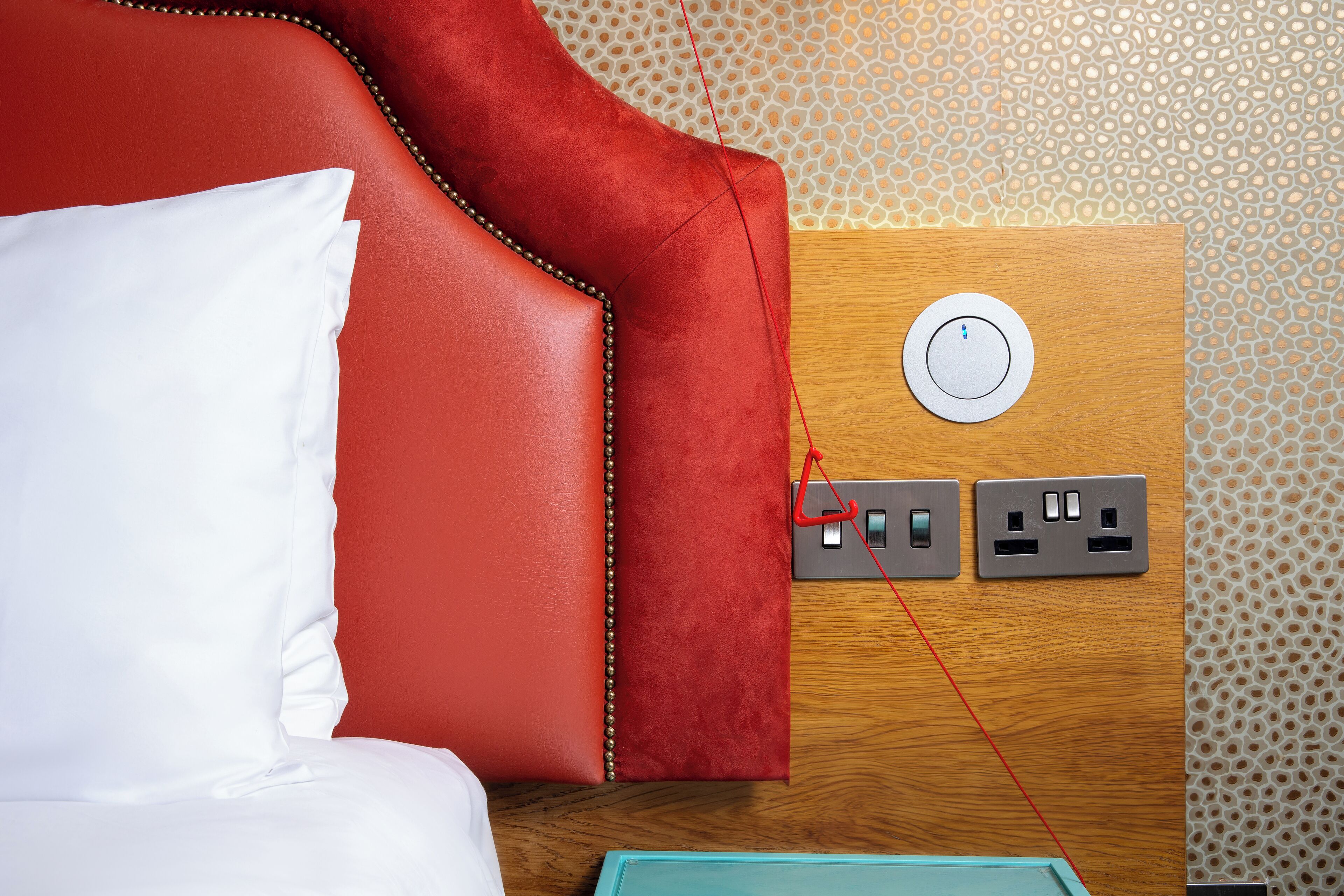 A red upholstered headboard with a white pillow, a wooden panel with a power outlet, and a patterned wall.