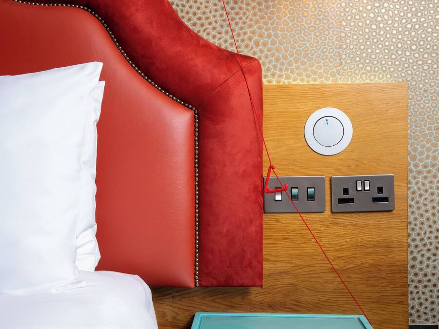 A red upholstered headboard with a white pillow, a wooden panel with a power outlet, and a patterned wall.