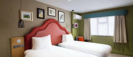 Ein Hotelzimmer mit zwei Betten, einem roten Kopfteil, einem Fenster mit Vorhängen und einer Wandklimaanlage.