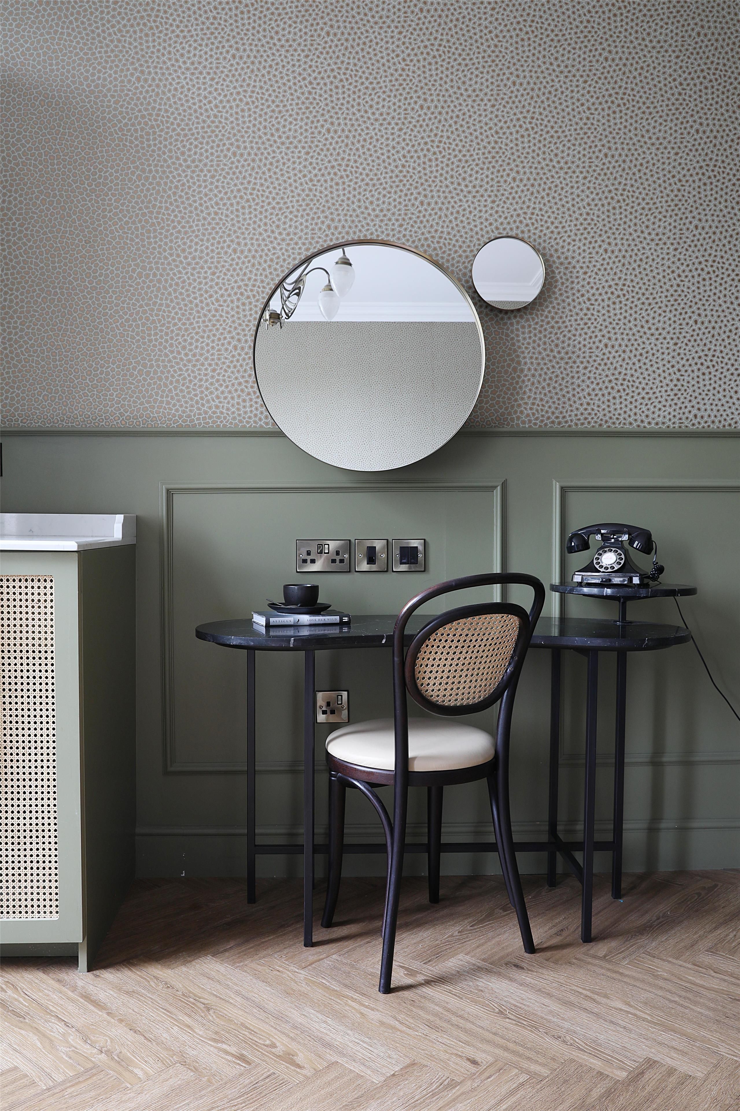 A room with a round mirror, a small table with a chair, a vintage telephone, and a wall-mounted light fixture.