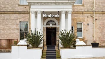 Ein Backsteinhaus mit weißem Eingang und dem Namen 'Elmbank' über der Tür. Auf beiden Seiten des Eingangs stehen je ein Blumentopf.