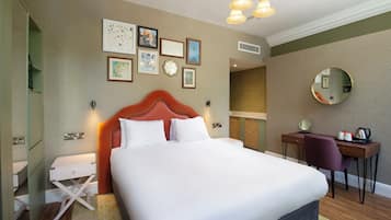 Een hotelkamer met een groot bed, een bureau met stoel, een spiegel en verschillende lijstjes met foto’s aan de muur.