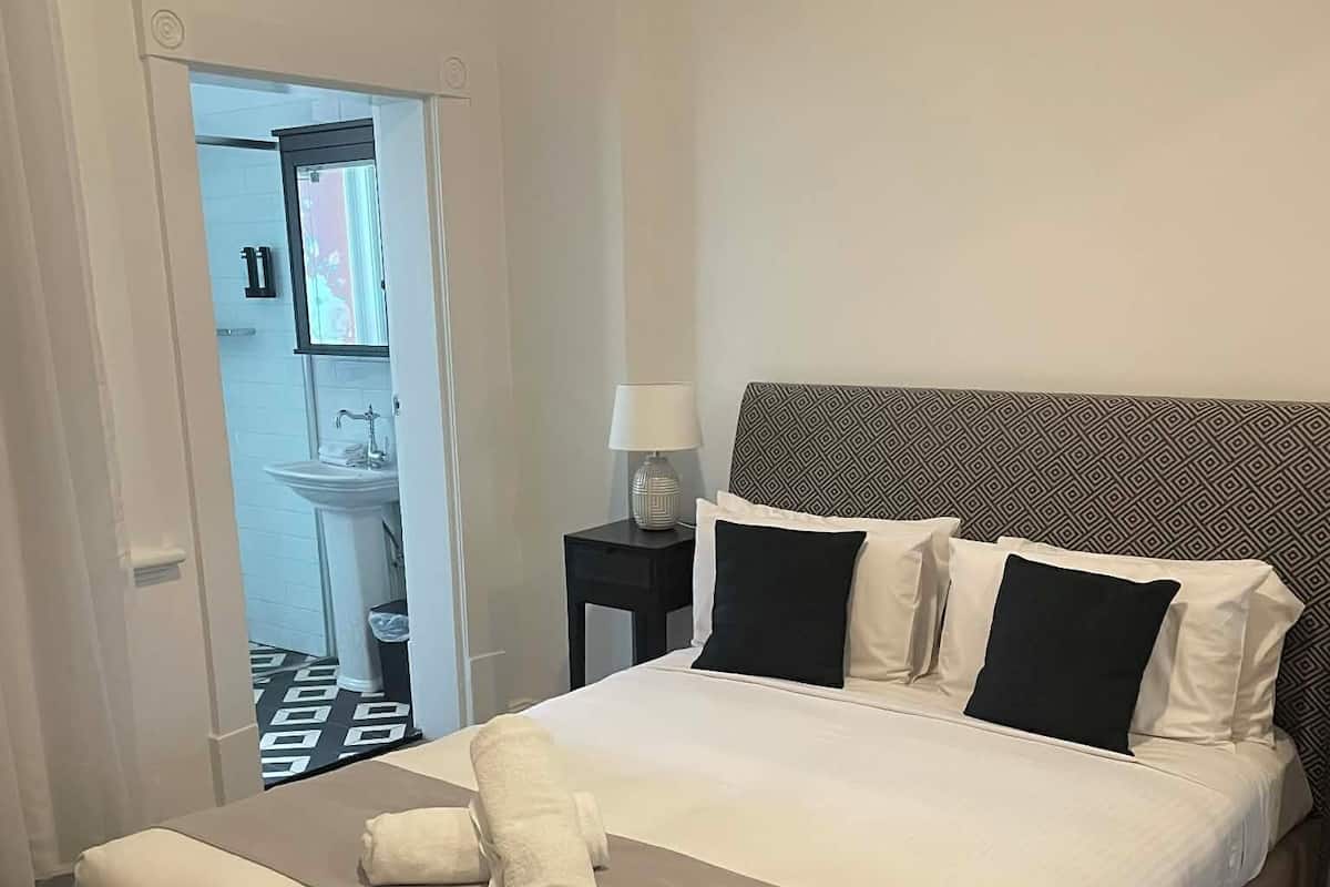 A hotel room with a large bed, a nightstand with a lamp, and a view of the bathroom through an open door.