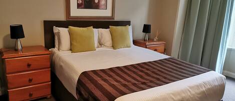 A hotel room with a large bed, two bedside tables, a nightstand, a framed artwork above the bed, and a window with curtains.