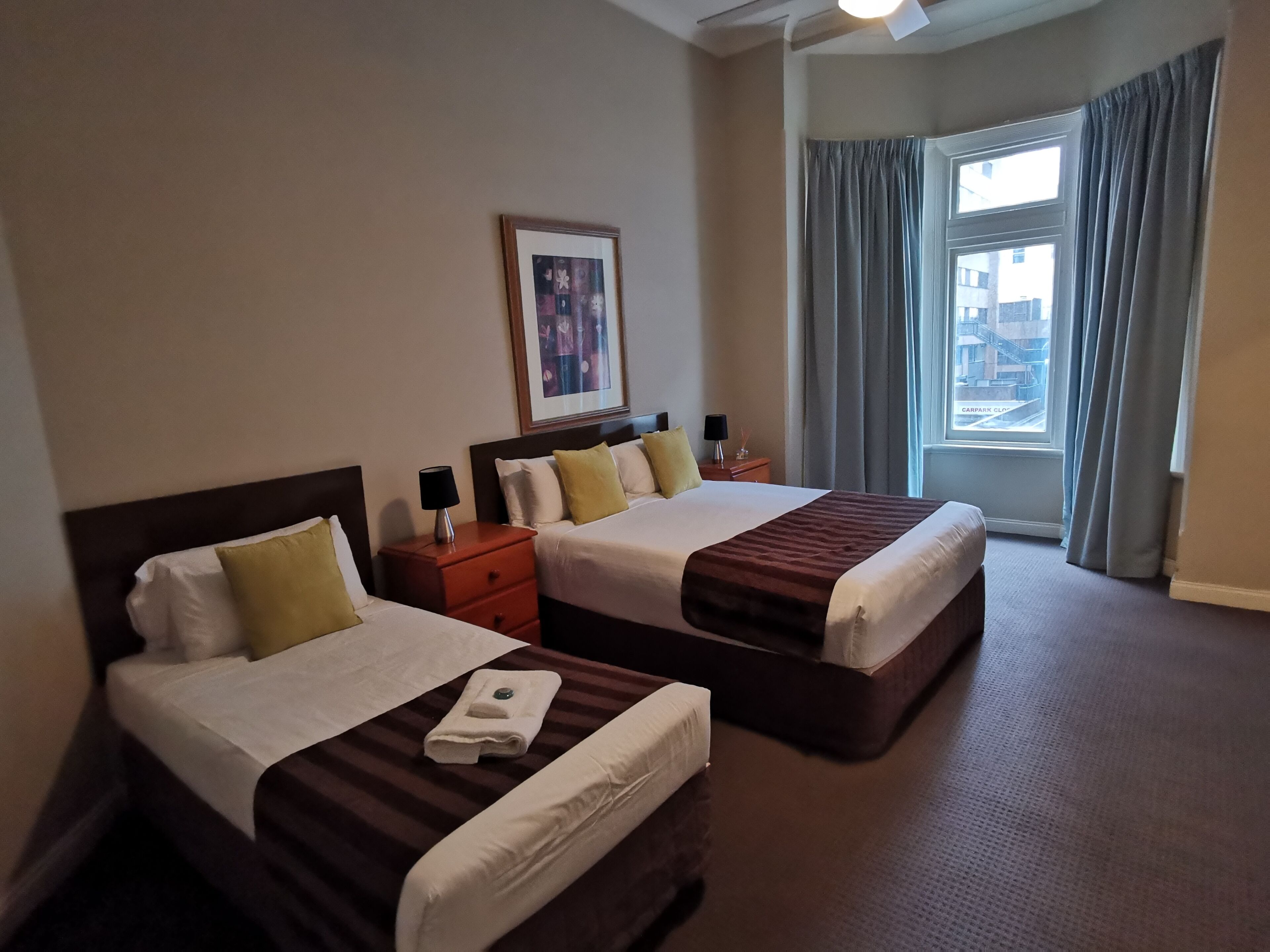 Habitación de hotel con dos camas, un cuadro en la pared, una mesita de noche con lámpara y una ventana con cortinas.