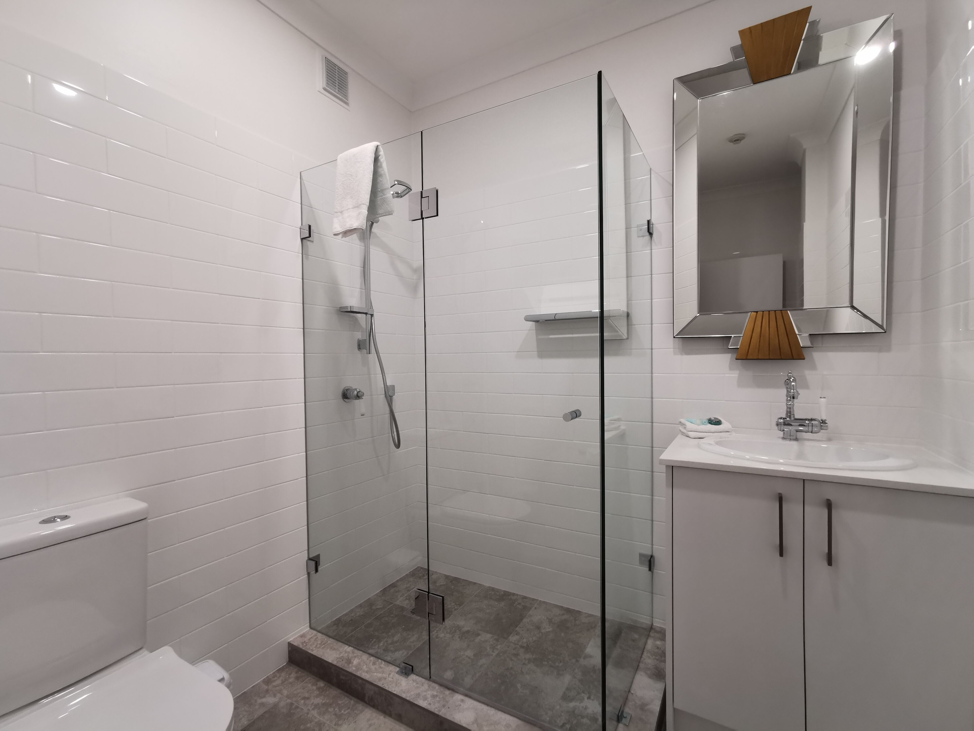 A modern bathroom with a glass-enclosed shower, white tiled walls, a white sink, and a mirror with a wooden frame.