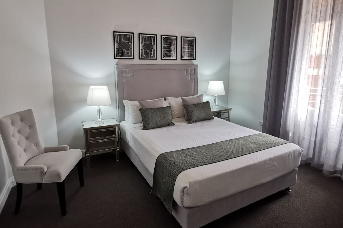 A neatly arranged hotel room with a large bed, two bedside tables with lamps, a chair, and a headboard with decorative panels.