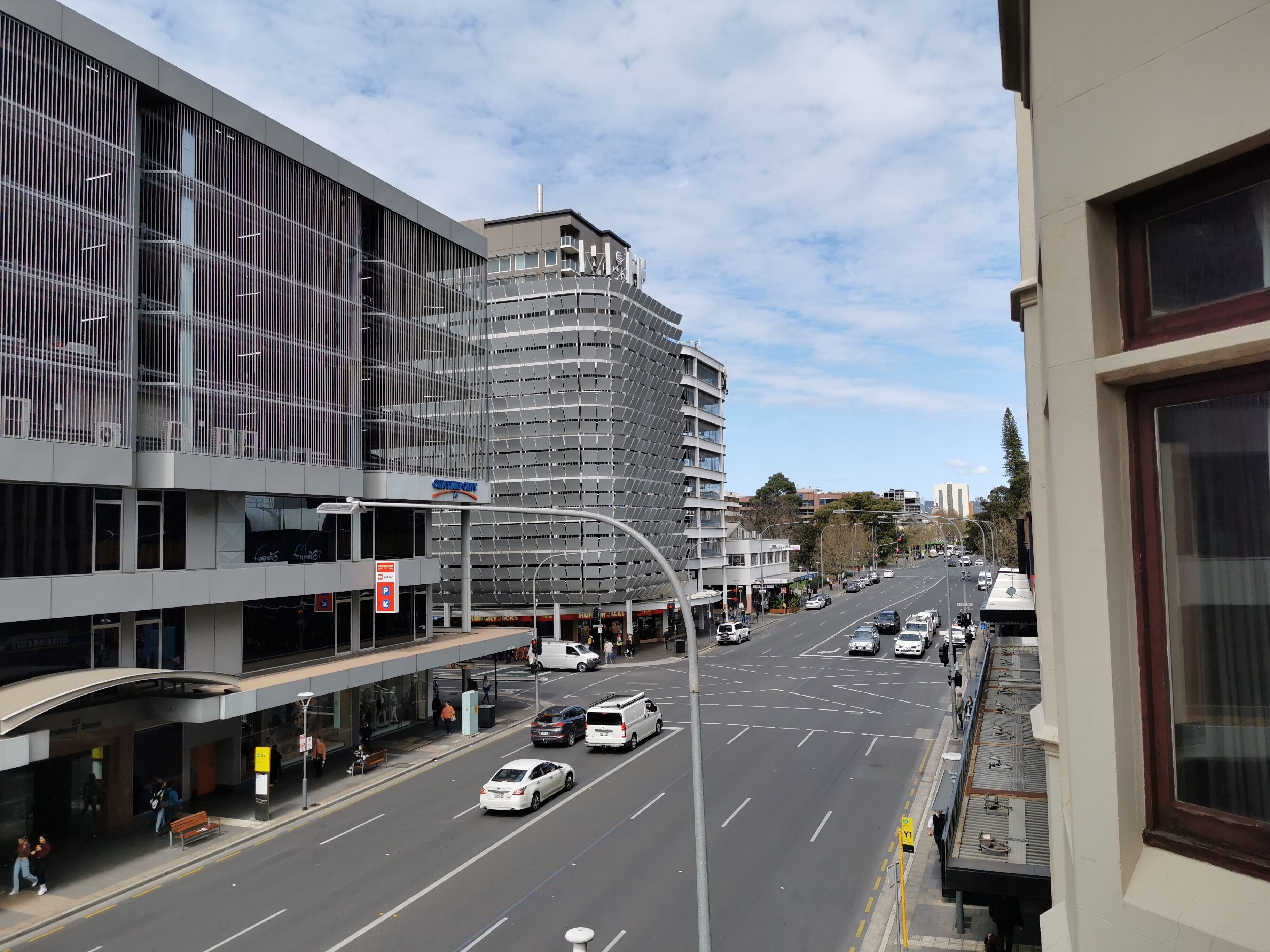 A modern urban street with multi-story buildings, a clear sky, and a variety of vehicles.