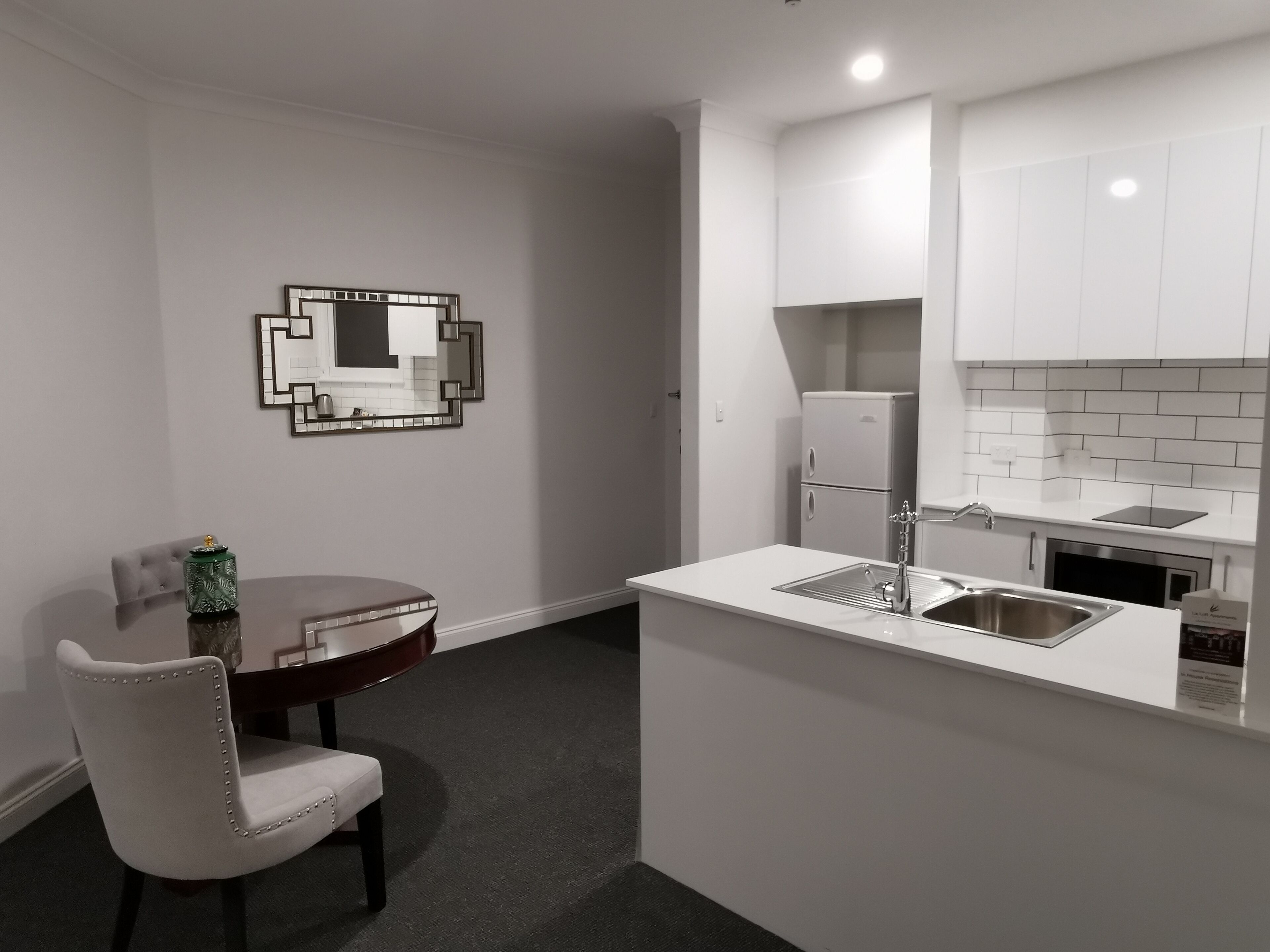 A compact kitchen with a white countertop, a sink, and a small refrigerator. There is a dining table with a chair and a decorative mirror on the wall.