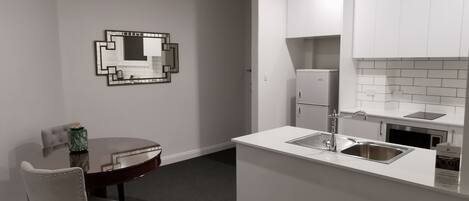 A compact kitchen with a white countertop, a sink, and a small refrigerator. There is a dining table with a chair and a decorative mirror on the wall.