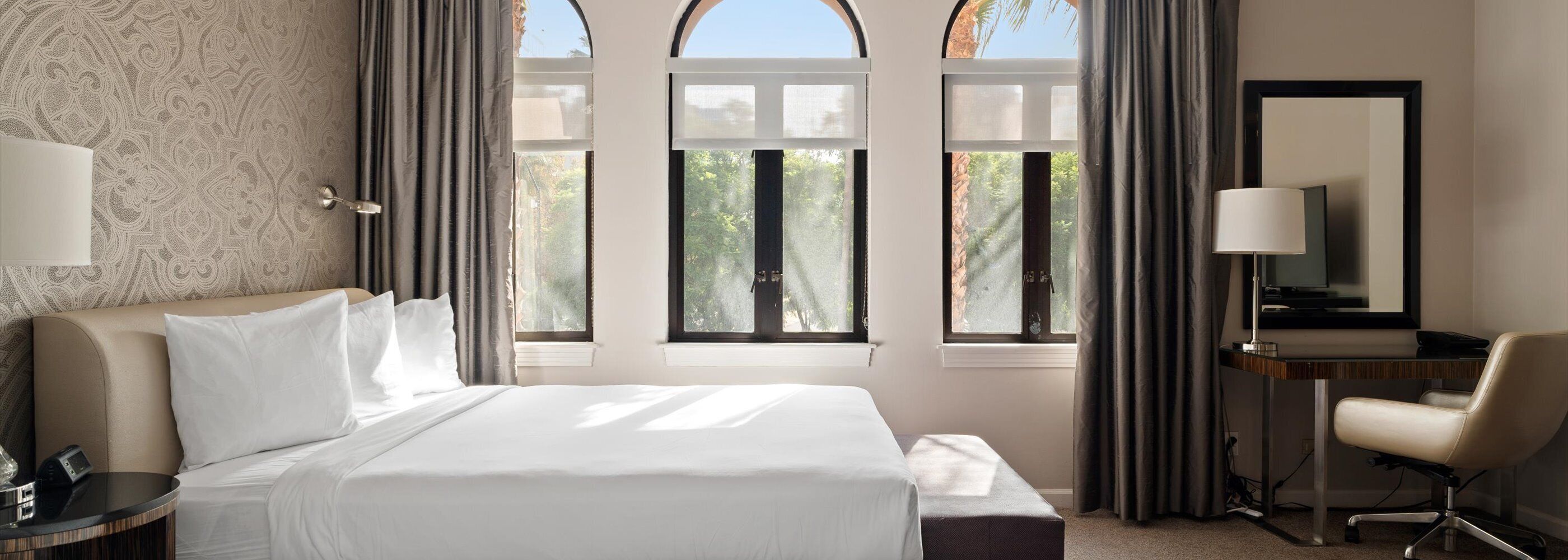 A hotel room with a bed, a desk, a chair, a lamp, and large windows with curtains.