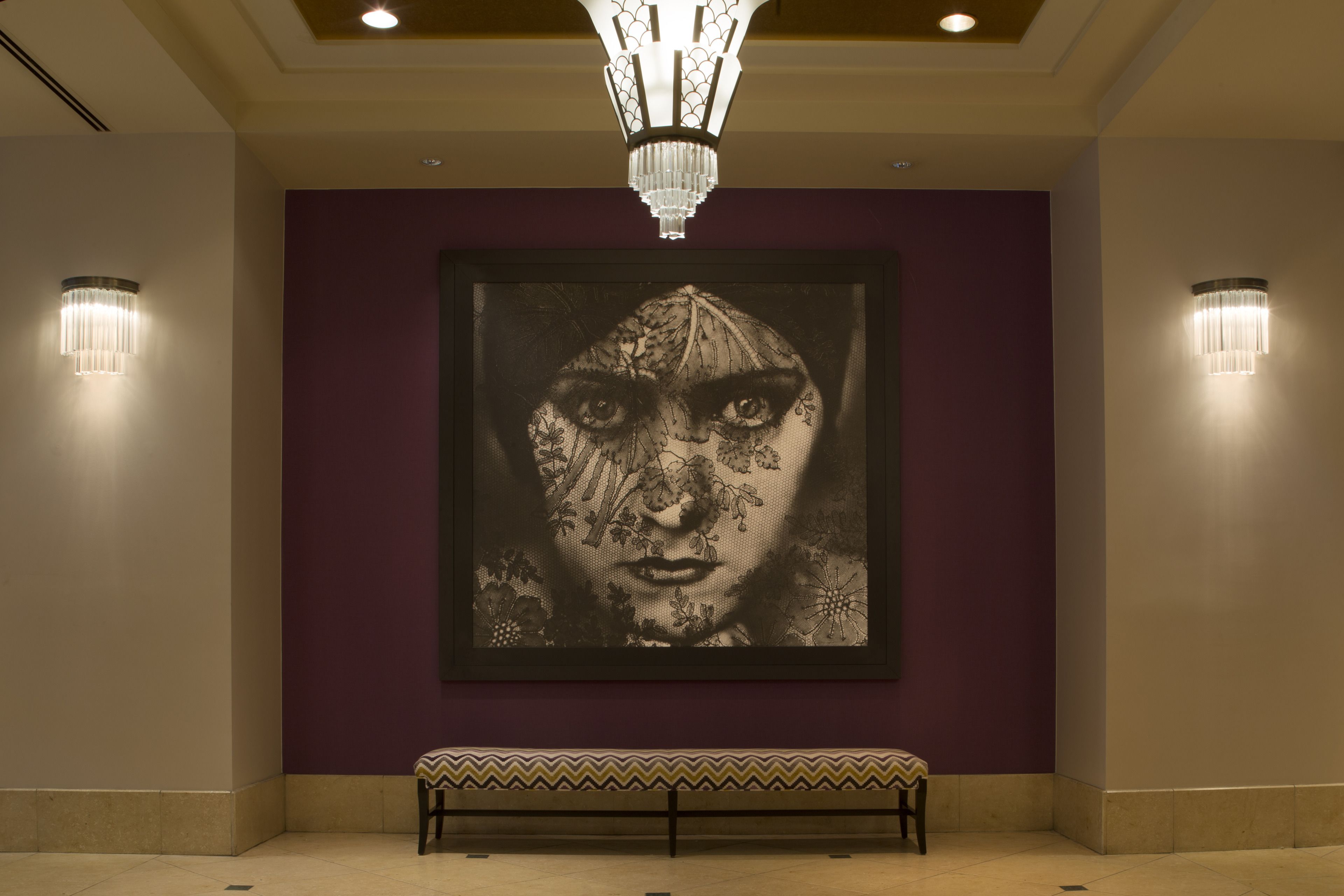 A lobby with a large framed artwork on the wall, a bench, and two wall-mounted light fixtures.
