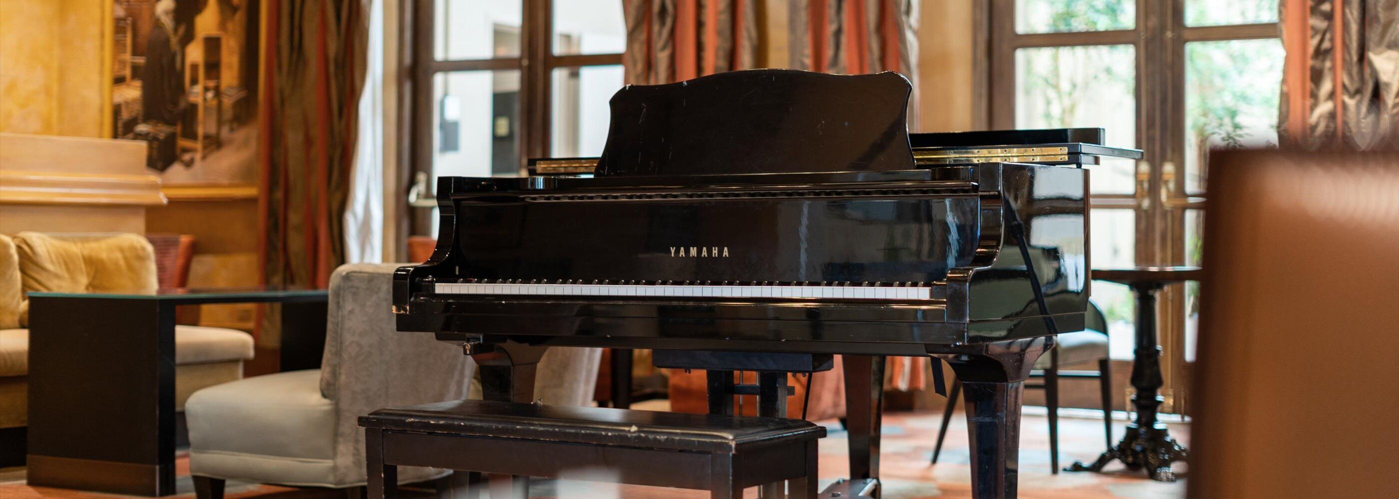 Un pianoforte a coda in una stanza luminosa, con ampie finestre, un divano e un tavolo.
