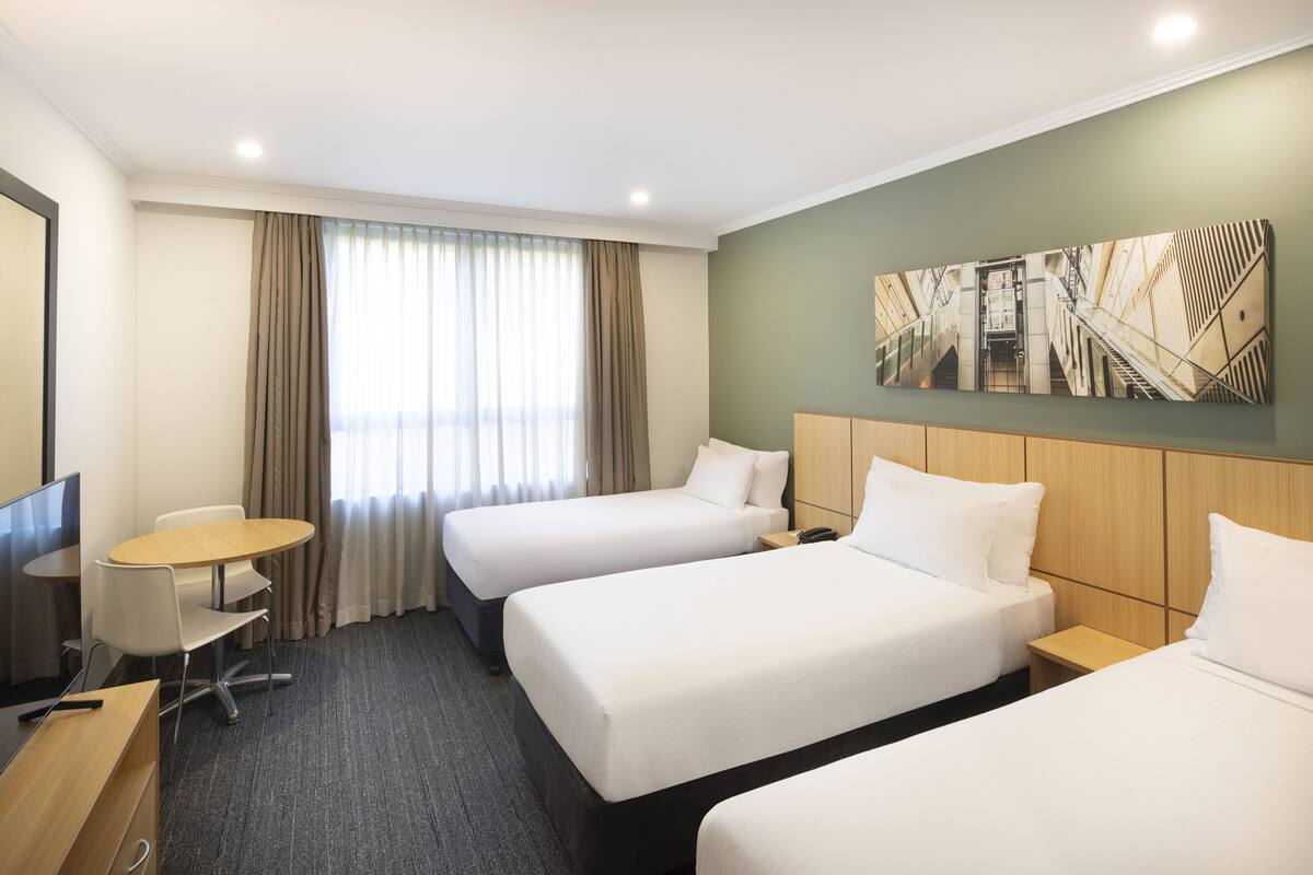 Ein Hotelzimmer mit zwei Betten, einem Schreibtisch, einem Stuhl, einem Fernseher und einem großen Fenster mit Vorhängen.