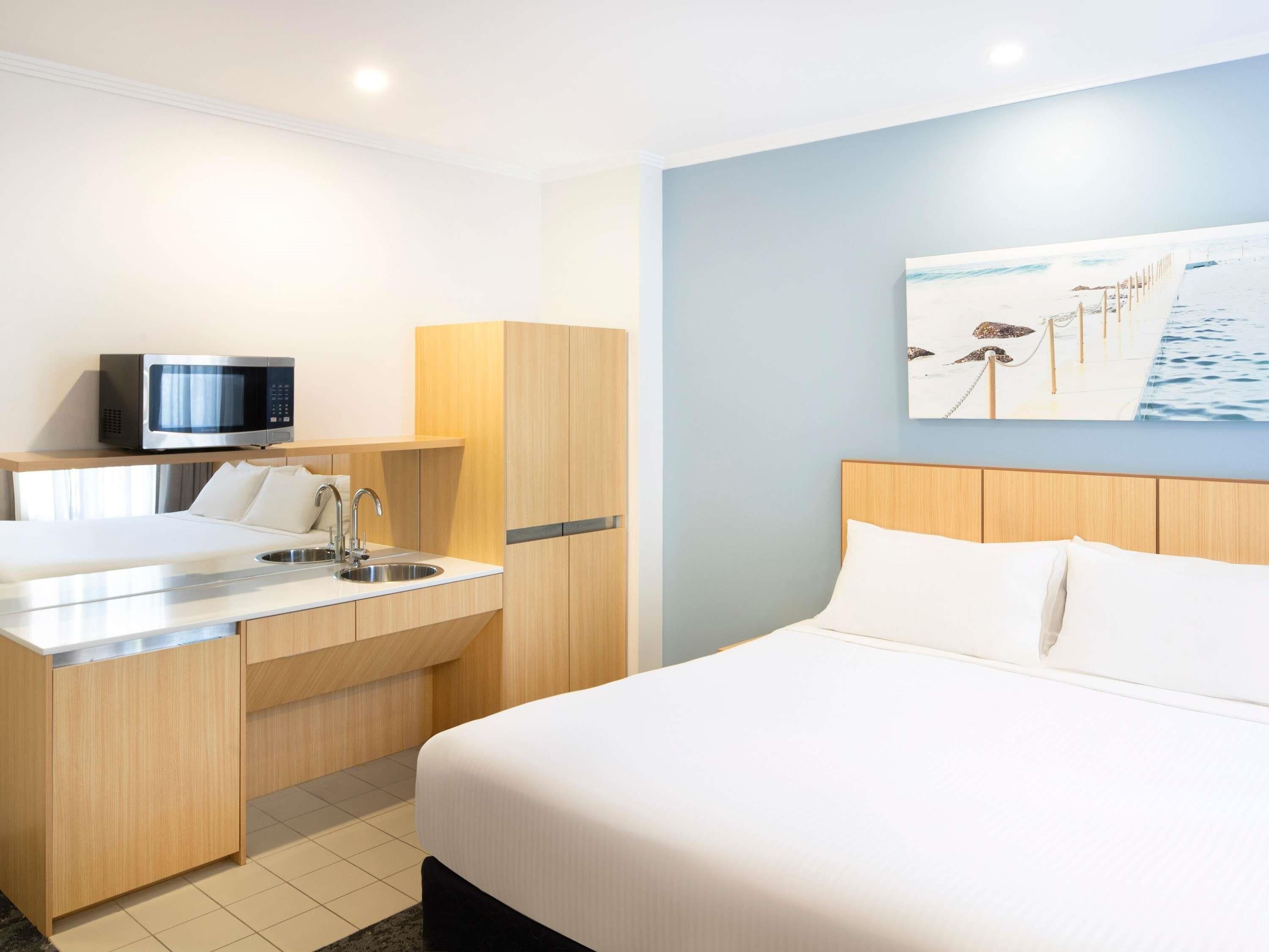 A hotel room with a bed, a kitchenette with a microwave, a sink, and a painting of a beach scene.