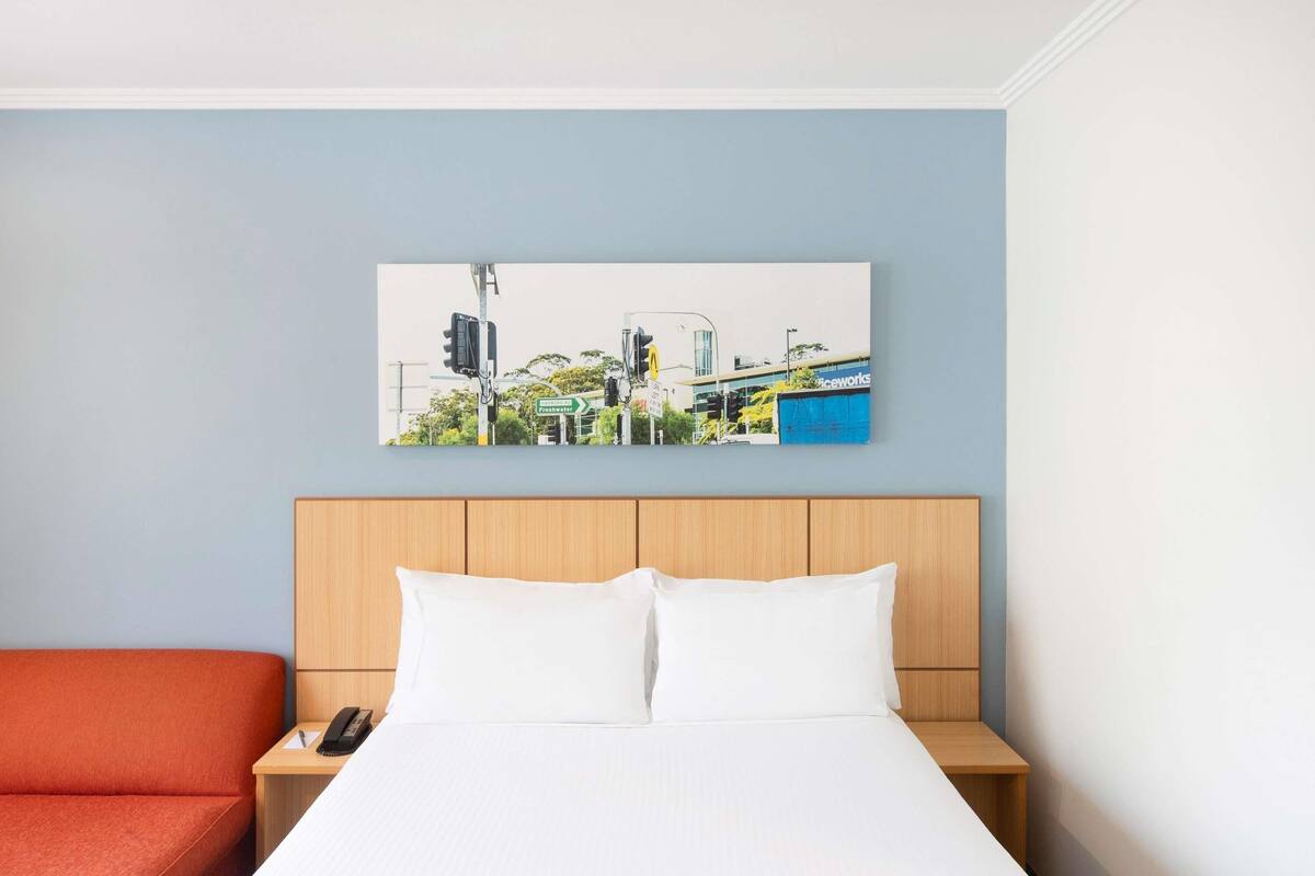 A hotel room with a bed, a nightstand, a red chair, and a cityscape painting on the wall.