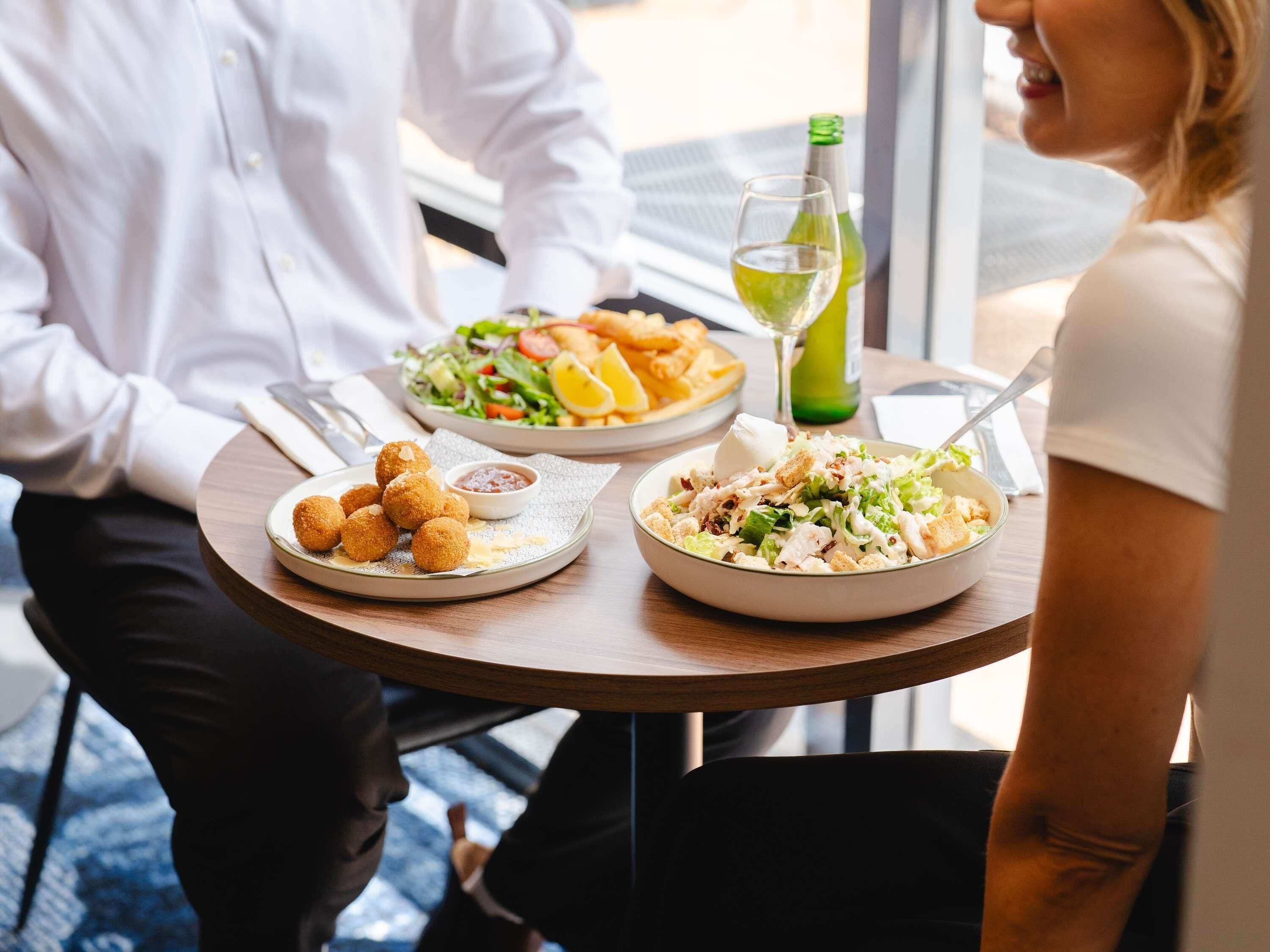 A dining table with two plates of food, a bottle of green drink, and a glass of white wine.