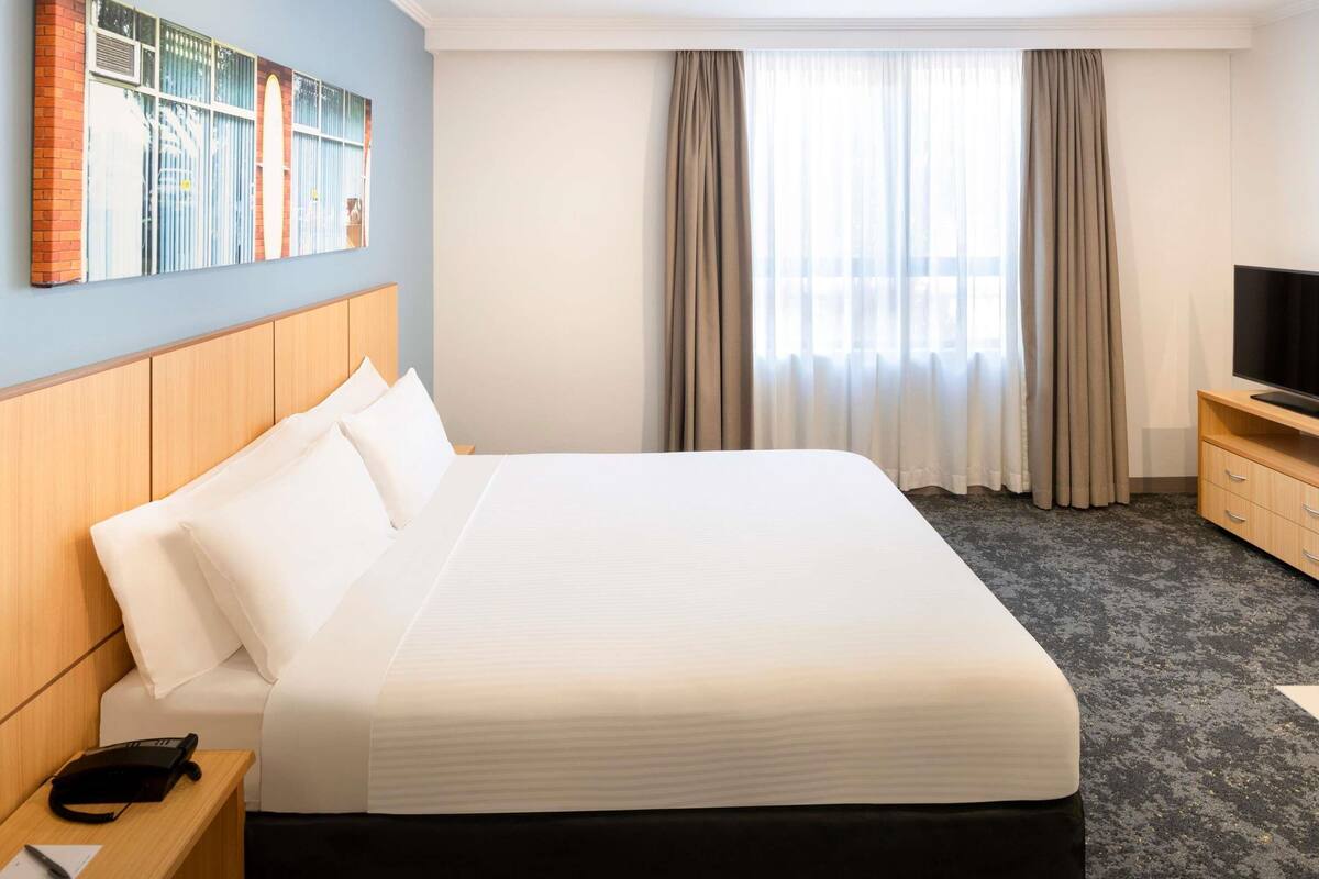 A hotel room with a large bed, a television on a wooden stand, a desk with a telephone, and a painting on the wall.