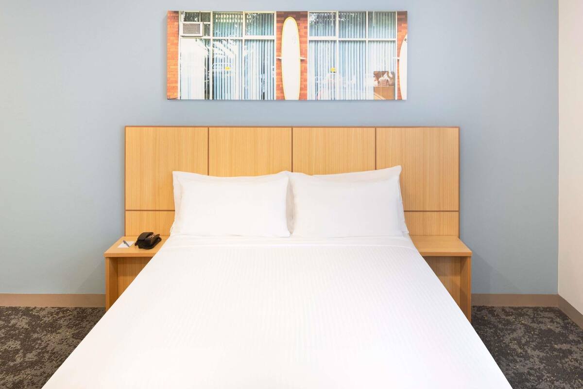 A hotel room with a wooden headboard, a large bed with white linens, a nightstand with a phone, and a wall-mounted artwork.