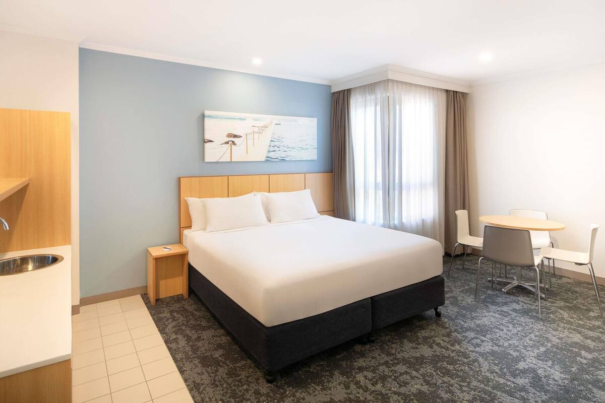 A hotel room with a bed, a desk, a chair, a painting on the wall, and a window with curtains.