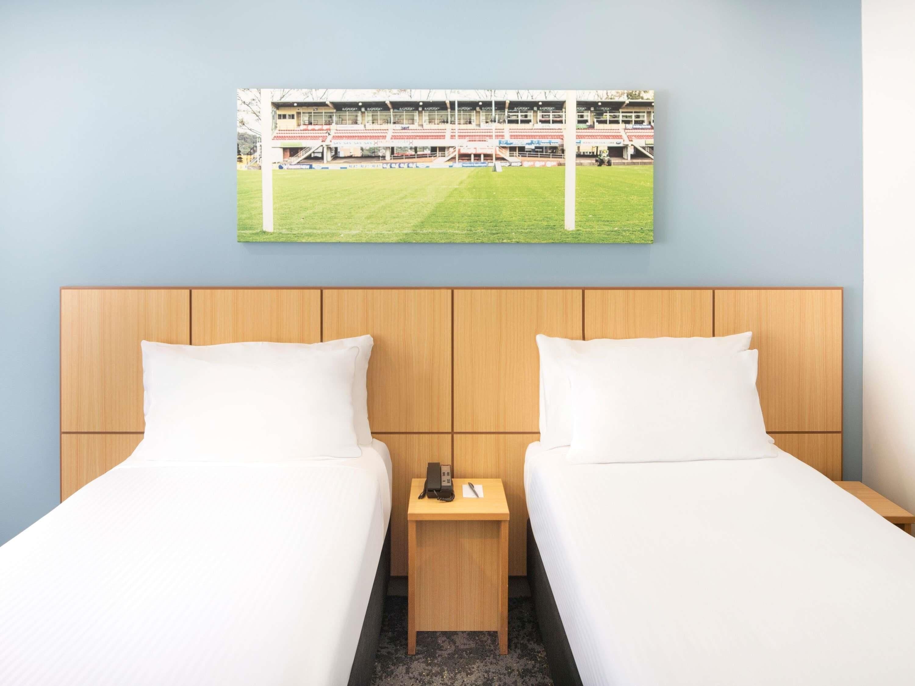 A hotel room with two beds, a wooden headboard, a small bedside table with a phone, and a framed picture of a stadium on the wall.