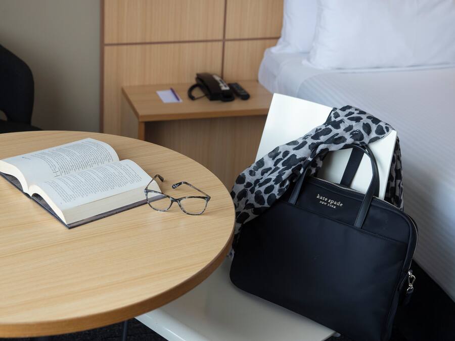 A hotel room with a round wooden table, a book, glasses, a black handbag, a bed, and a nightstand with a phone and a notepad.