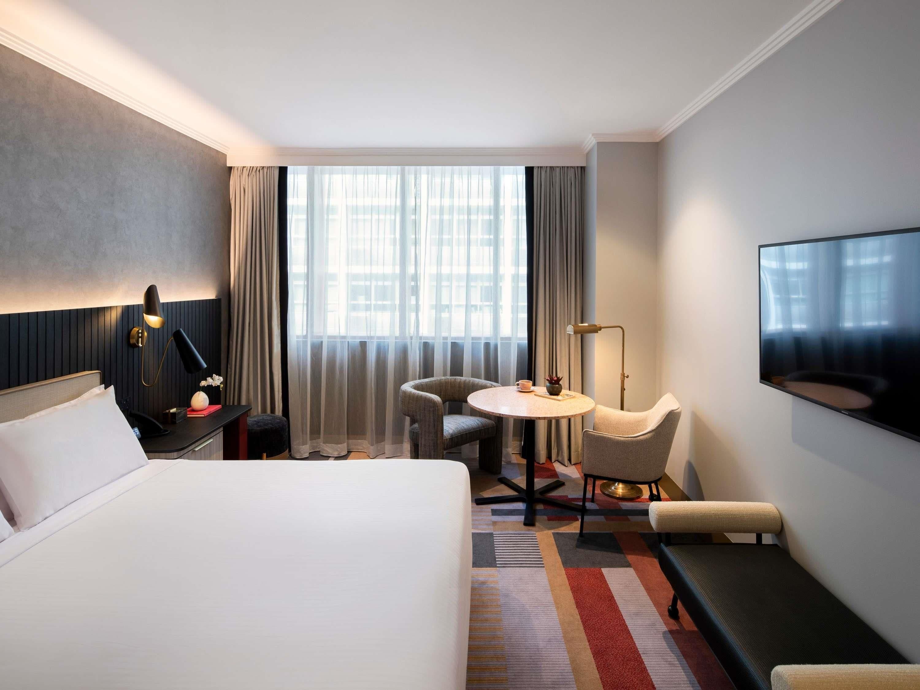 Een hotelkamer met een bed, een bureau met lamp, een ronde tafel met stoelen, een televisie en een raam met gordijnen.