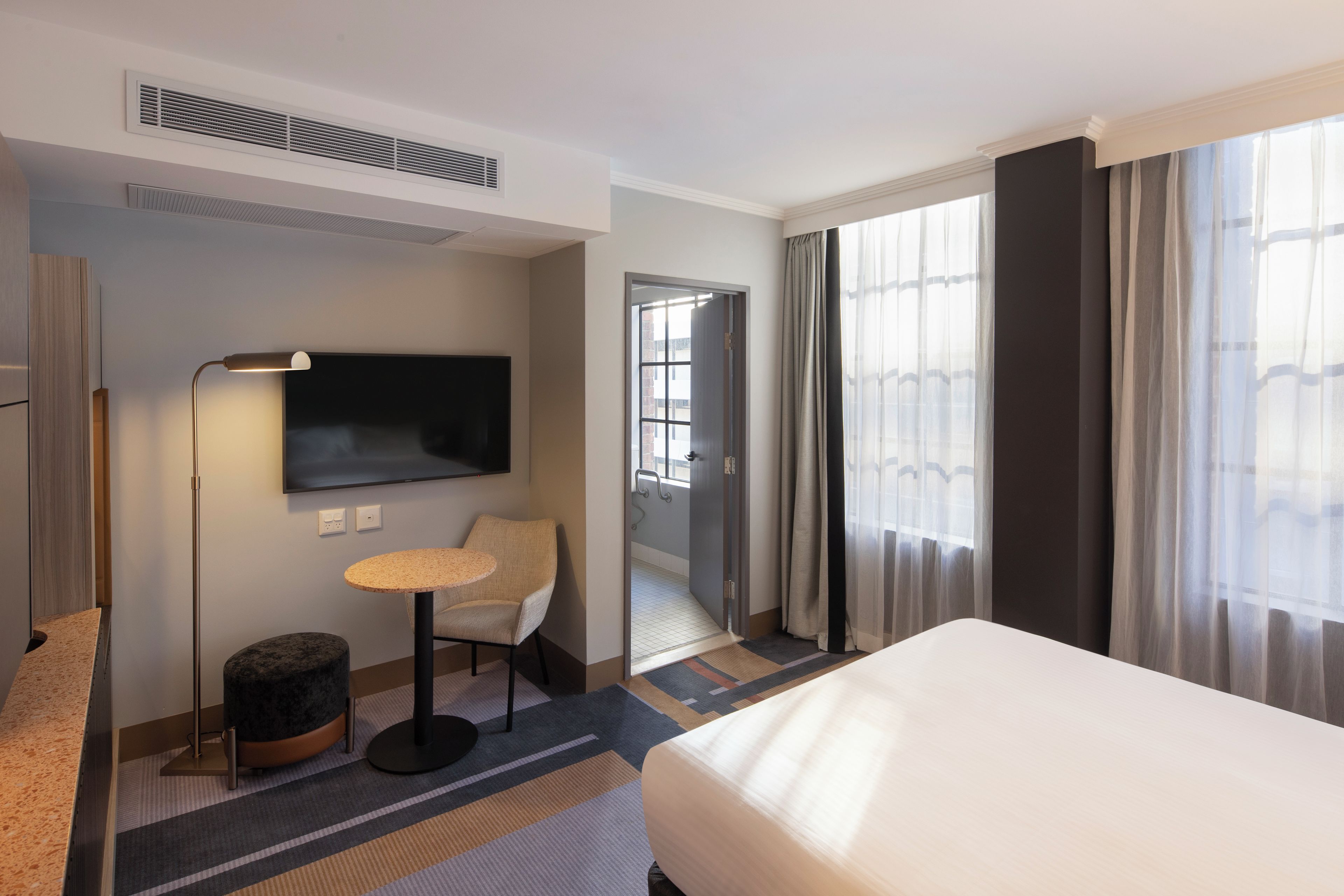 A hotel room with a bed, a television, a small table, a chair, a lamp, and a view of the outside through the window.