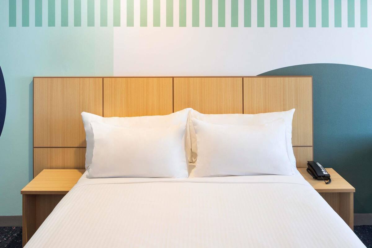 A hotel room with a bed, two pillows, a wooden headboard, a bedside table with a phone, and a wall with a geometric design.