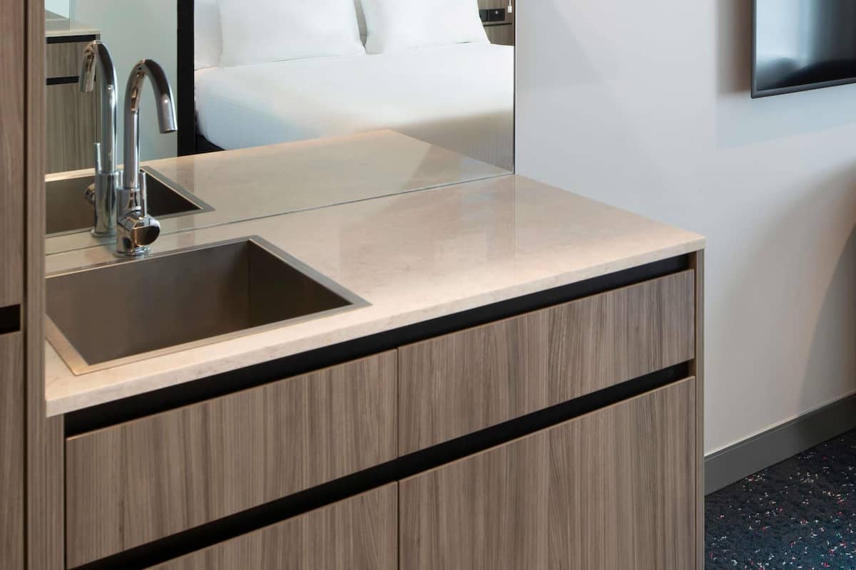 A modern bathroom with a sink, mirror, and a bed visible in the background.