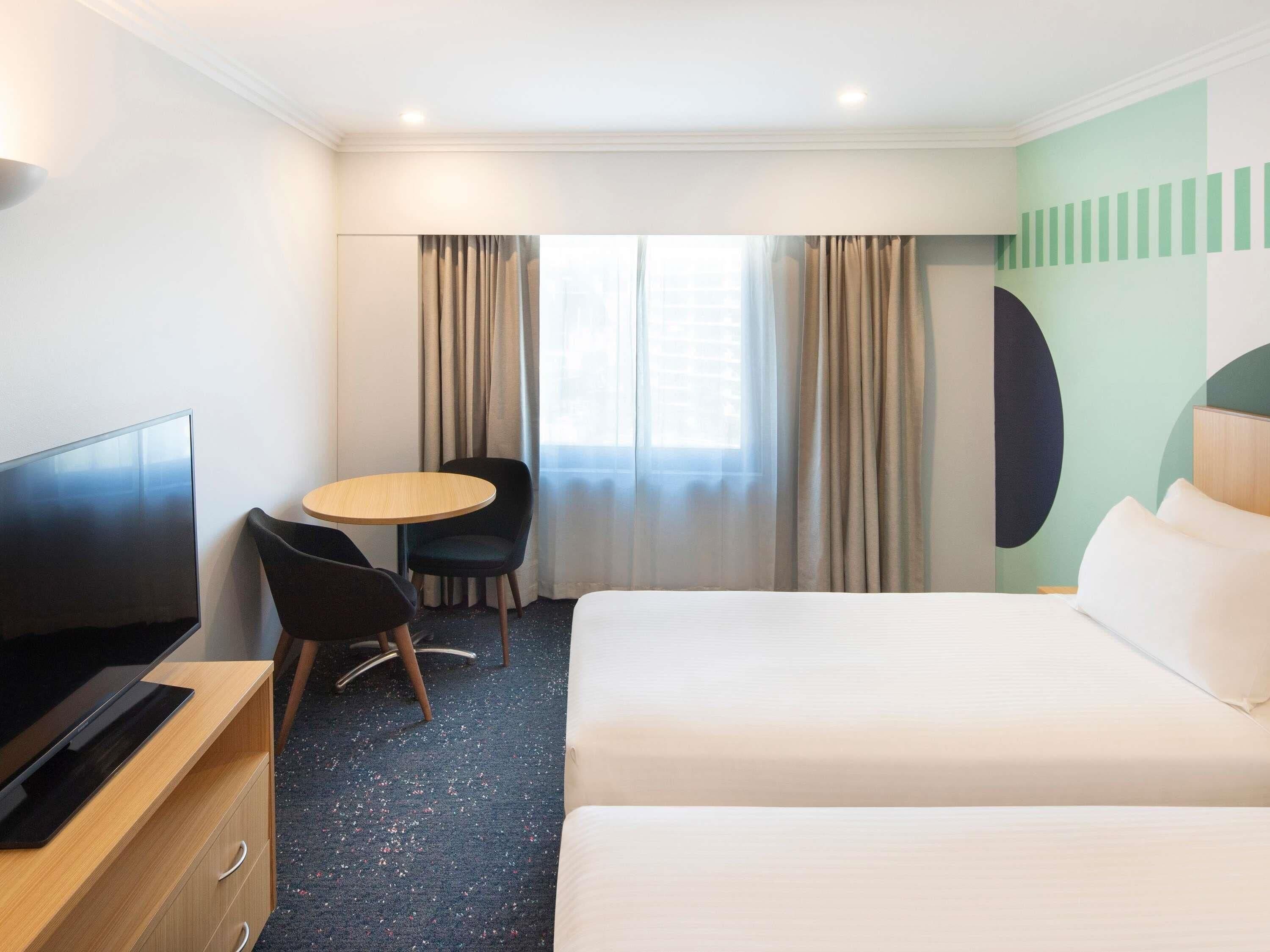 A hotel room with a bed, a TV, a desk, a chair, and a window with curtains.