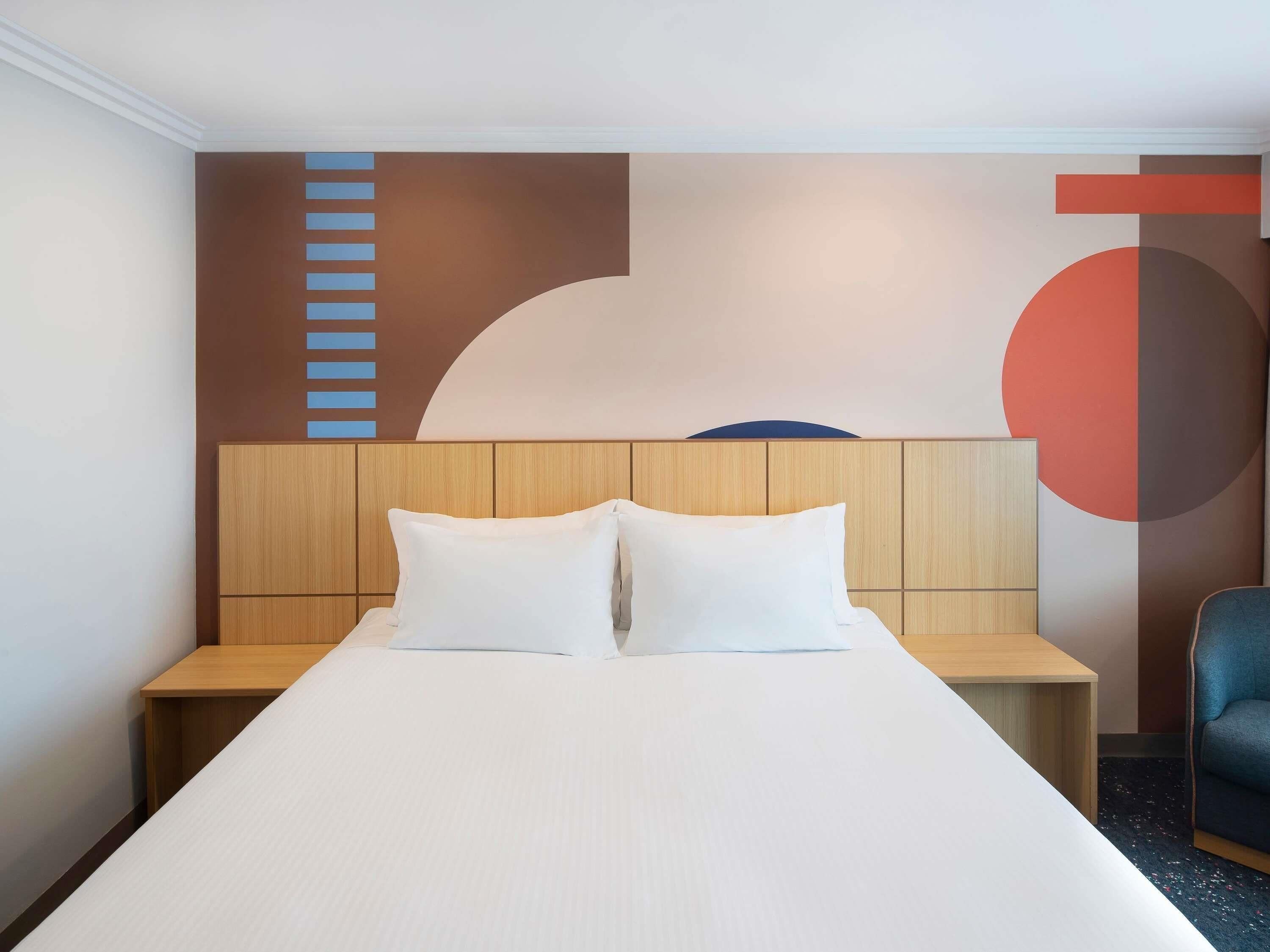 A hotel room with a bed, wooden headboard, two bedside tables, a chair, and a wall with abstract art.