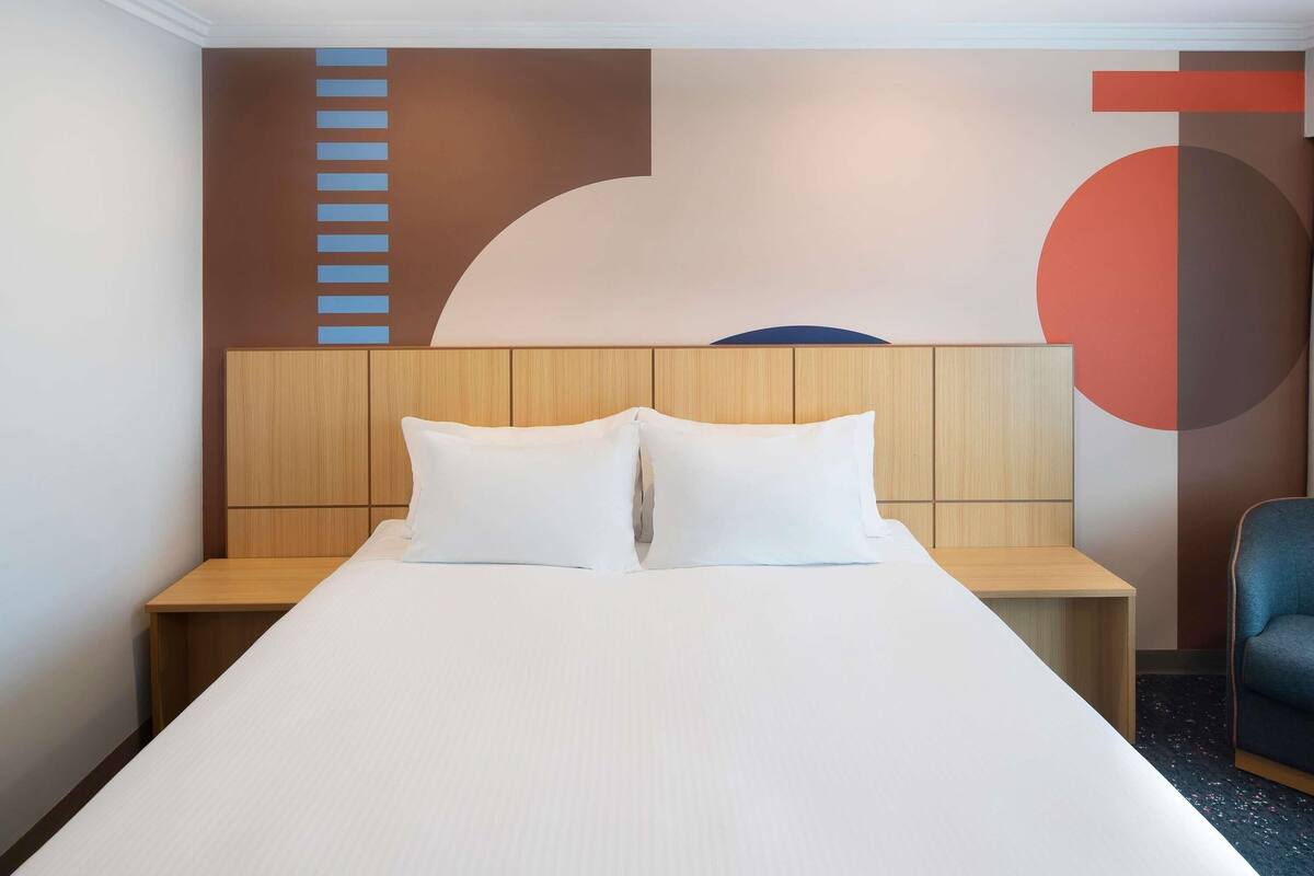 A hotel room with a bed, wooden headboard, two bedside tables, a chair, and a wall with abstract art.