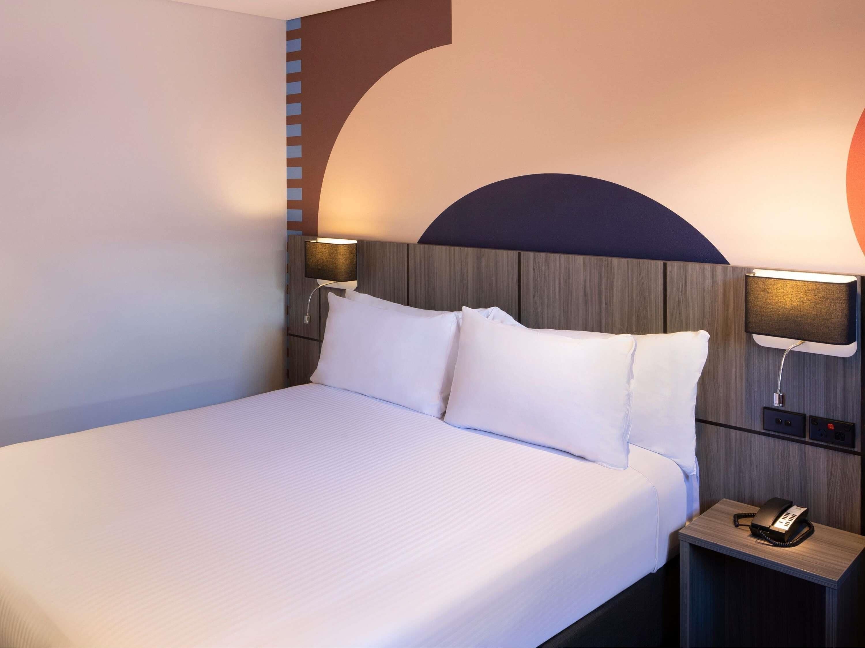 A hotel room with a large bed, two bedside lamps, a nightstand with a telephone, and a wall-mounted light.