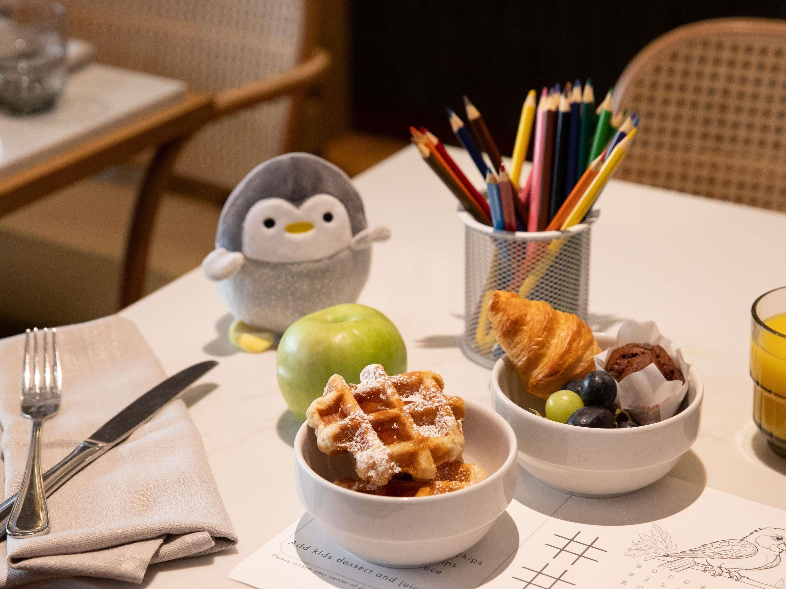 Un desayuno con waffles, croissants y fruta, un juguete de pingüino y una taza de lápices de colores.
