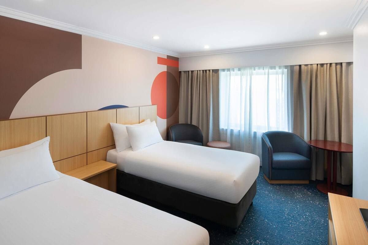 A hotel room with two beds, a small table, a chair, and a large window with curtains.