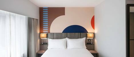 A hotel room with a large bed, two bedside tables with lamps, a headboard with a geometric design, and a partial view of a window with curtains.