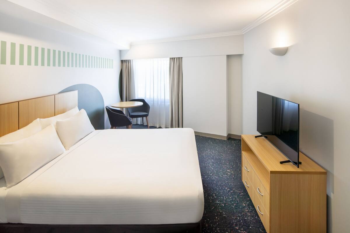 A hotel room with a large bed, a TV on a wooden stand, a small round table with two chairs, and a window with curtains.
