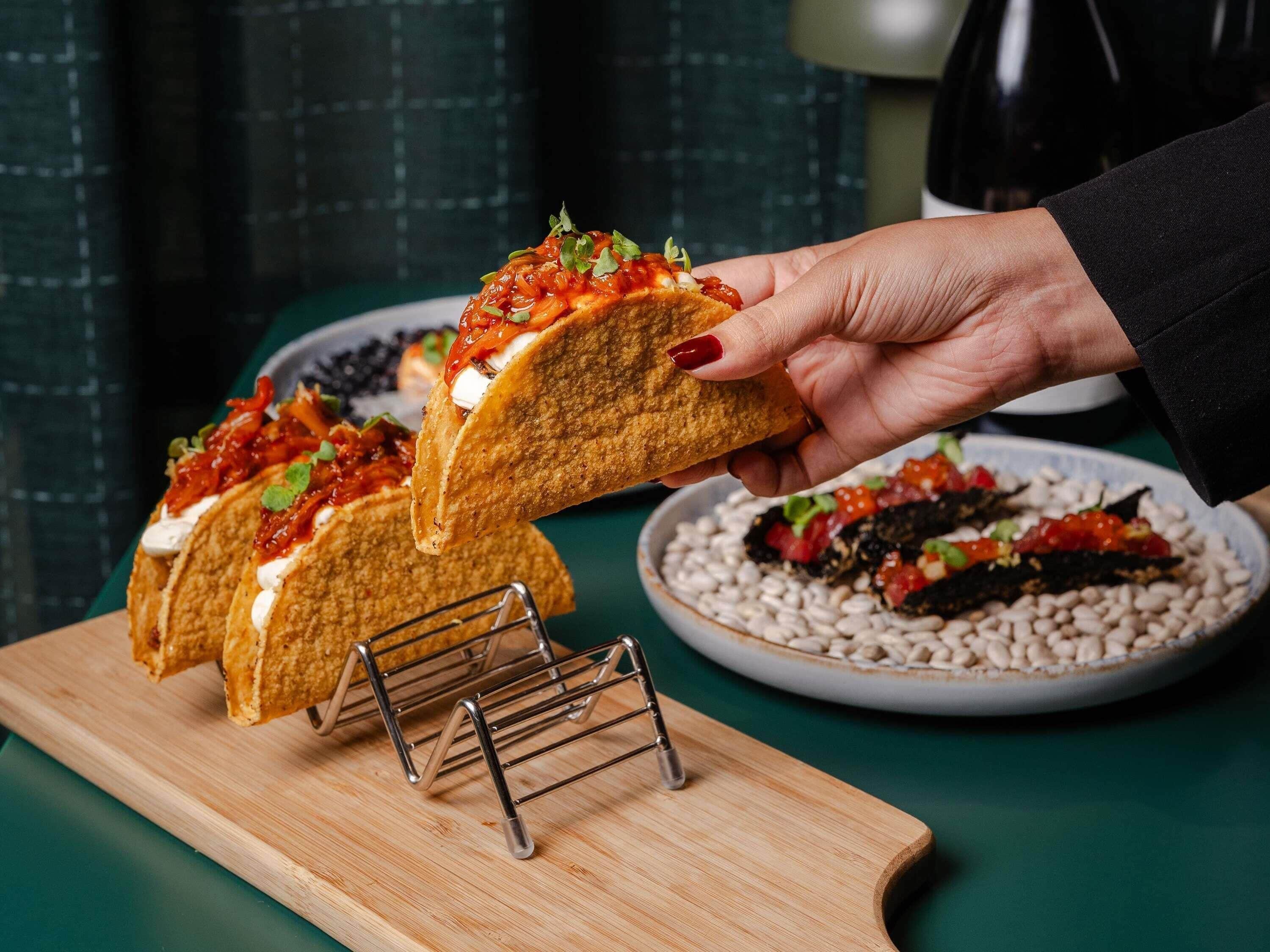 A hand holding a taco with toppings, a bowl of beans, and a metal rack on a wooden board.