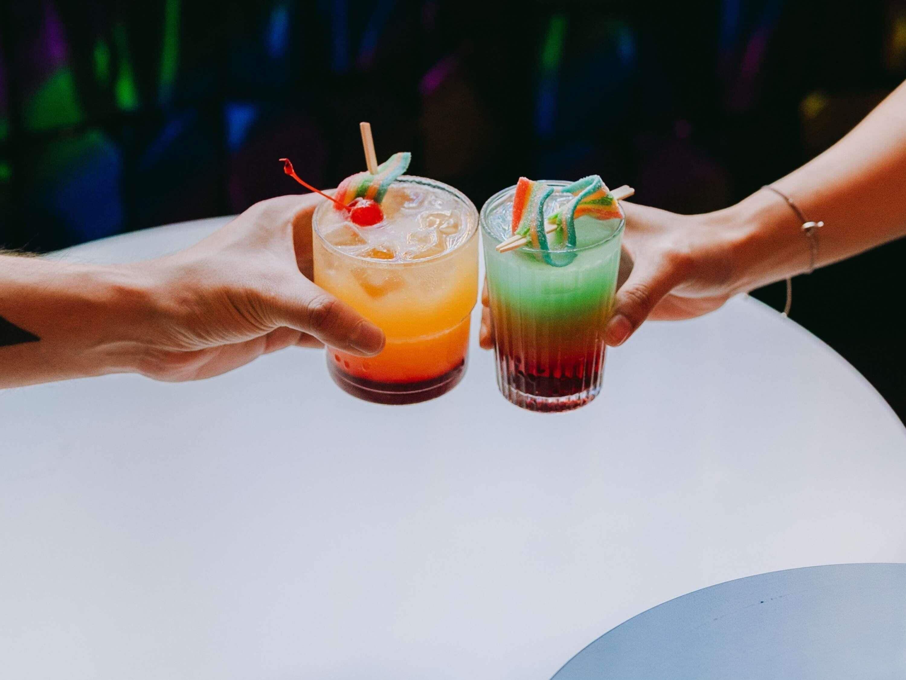 Two hands holding glasses of colorful cocktails with straws and garnishes.