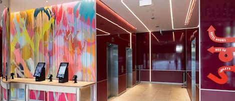 A hotel reception area with a colorful abstract wall, a reception desk with digital displays, and directional signs for hotel rooms, the restroom, and the elevator.